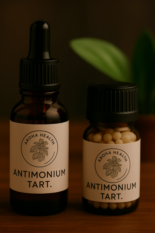 Two amber glass bottles of Aroha Health remedies labeled “Antimonium tart.” are placed on a wooden surface. The larger bottle with a black dropper cap contains liquid, and the smaller bottle holds round beige pellets. Both labels display the Aroha Health logo with a botanical illustration, with a blurred green plant in the background.