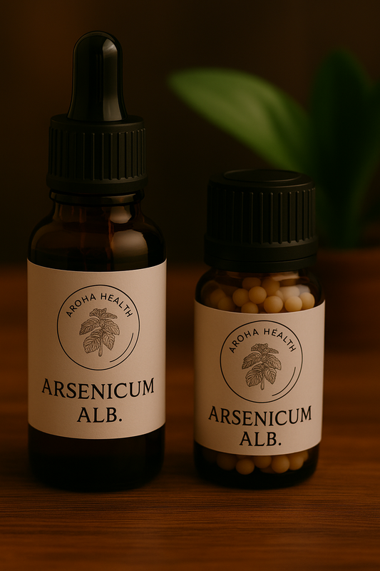 Two amber glass bottles of Aroha Health remedies labeled “Arsenicum alb.” stand on a wooden surface. The larger bottle with a black dropper cap is filled with liquid, while the smaller bottle contains round beige pellets. Both labels display the Aroha Health logo with a plant illustration, and a blurred green leaf is visible in the background.
